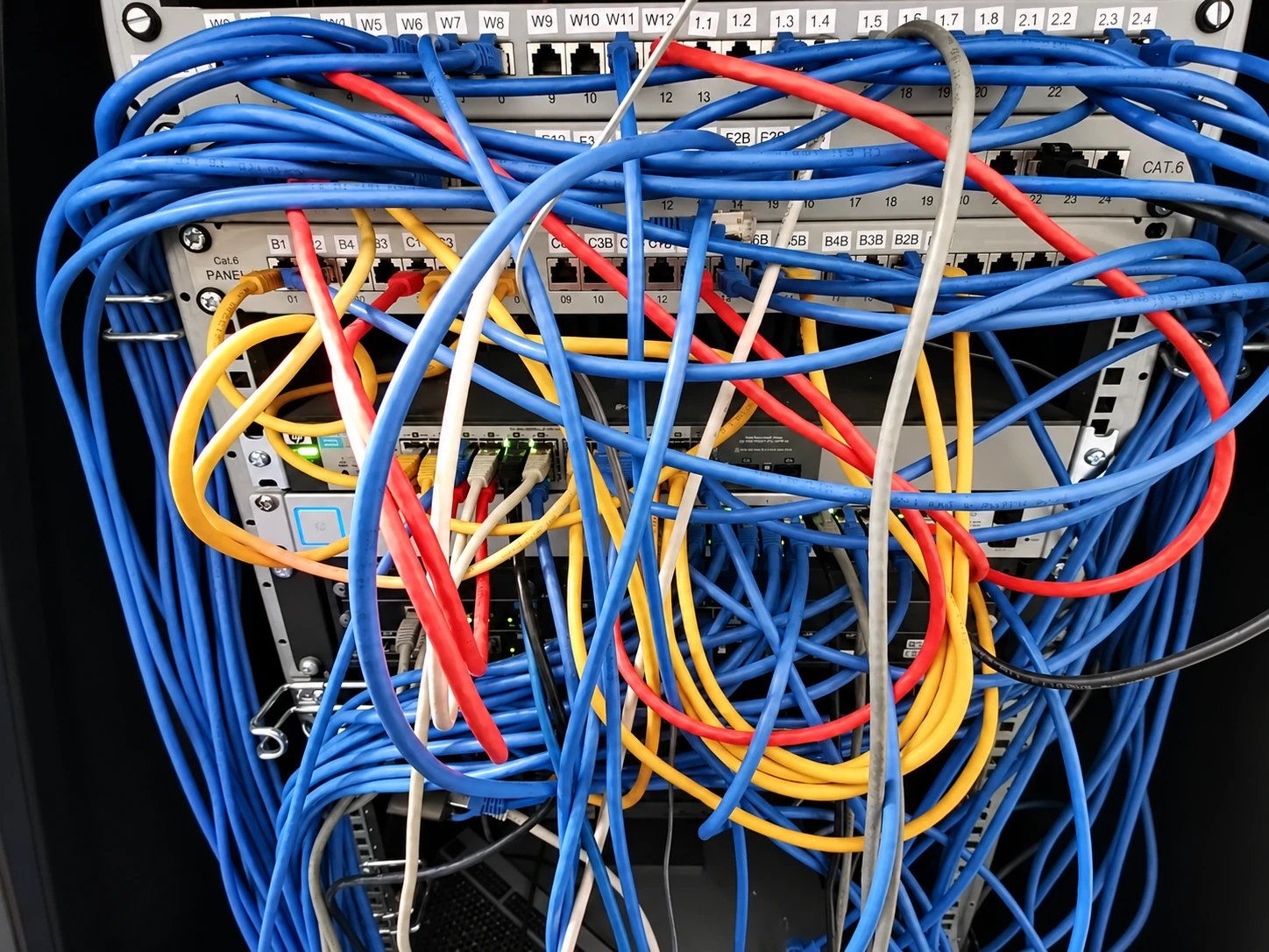 Netzwerkschrank vorher: lange blaue, rote, gelbe und graue Patchkabel hängen kreuz und quer über Patchfeldern und Switches.
